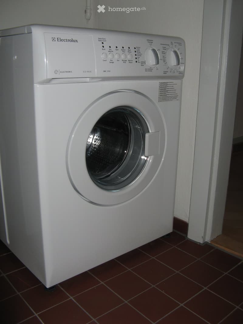 A white washing machine with a control panel is placed on a tiled floor in a bathroom.