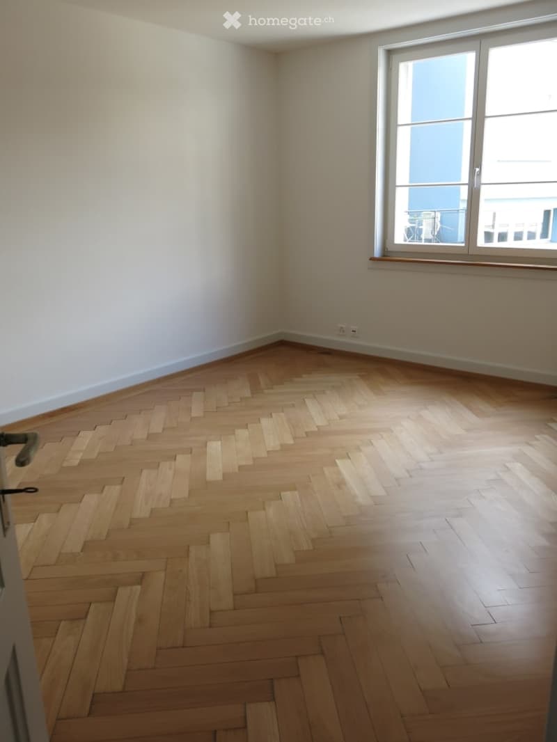 Empty room with wooden herringbone flooring and white walls, featuring a large window with a view of the outside.