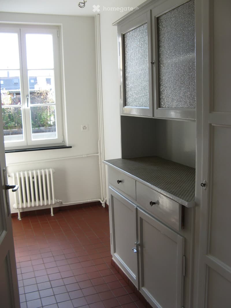Empty room with red tiled floor, grey cabinets, and a radiator near the window. The room has white walls and a window with a view of the outside.