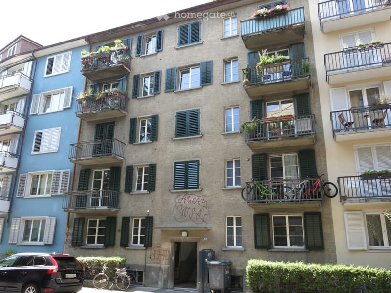 Multi-story residential building with multiple balconies, green shutters, and bicycles parked on one balcony. The building has a mix of light-colored and blue facades.