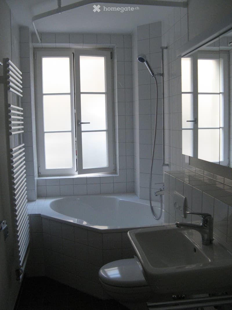 Modern bathroom with a bathtub, toilet, sink, showerhead, and radiator. The room features tiled walls and floors, large windows, and a mirror above the sink.