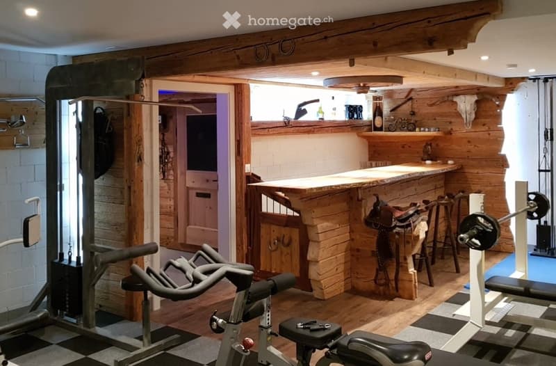 A rustic-themed room with a wooden bar counter, stools, a saddle, exercise bike, weight machine, and a shelf with a bottle and other items.