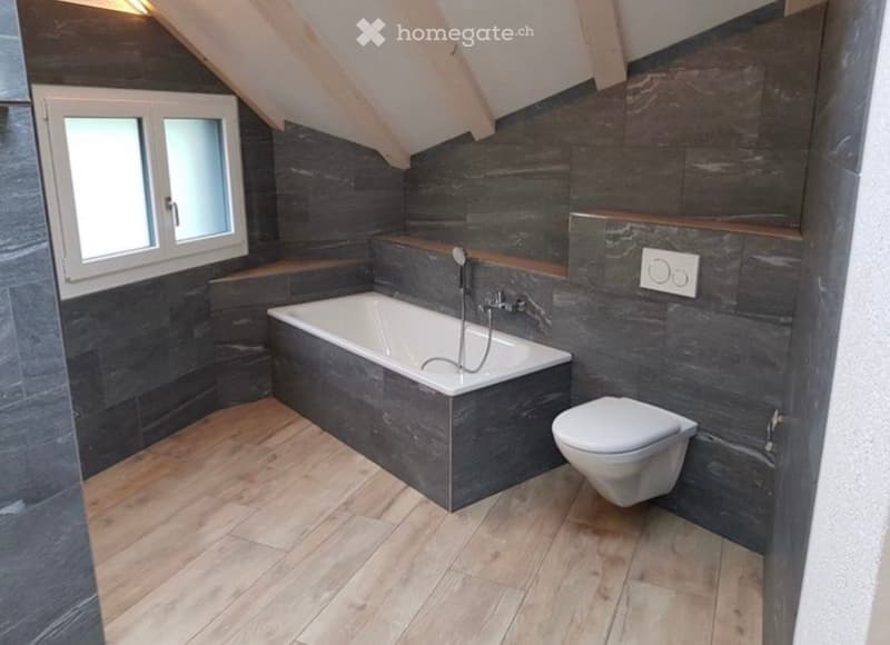 Modern bathroom with dark grey tiled walls, a white bathtub, a toilet, and a window with frosted glass. The floor is light wooden, and the space features a sleek, contemporary design.