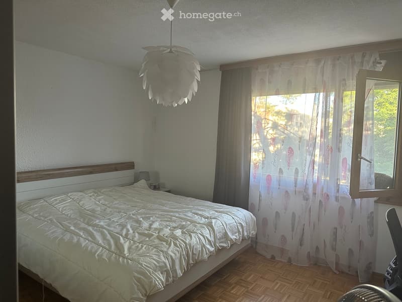 A well-lit bedroom with a large bed, wooden nightstand, and a decorative hanging light fixture. The room features wooden flooring and sheer curtains with a feather design.