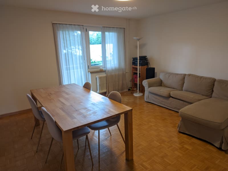 A modern living room with a wooden table, four chairs, a beige sofa, a floor lamp, and a small shelf with electronic devices.