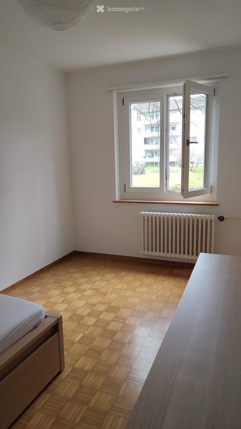 A furnished room with wooden flooring, a bed, a radiator, and a large window with a view of a residential area.