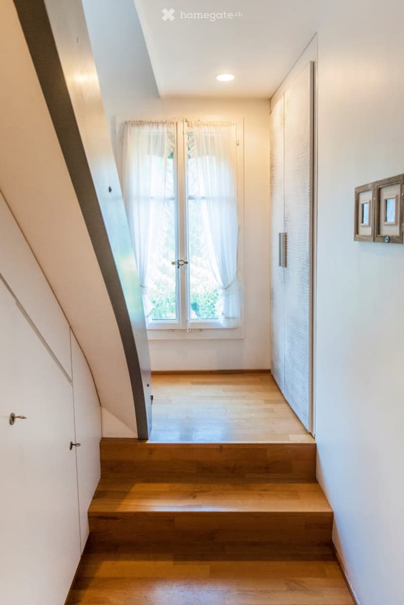 A modern, well-lit hallway with wooden flooring, white walls, and a large window with sheer curtains. The hallway features built-in storage units on one side and a staircase leading upwards.