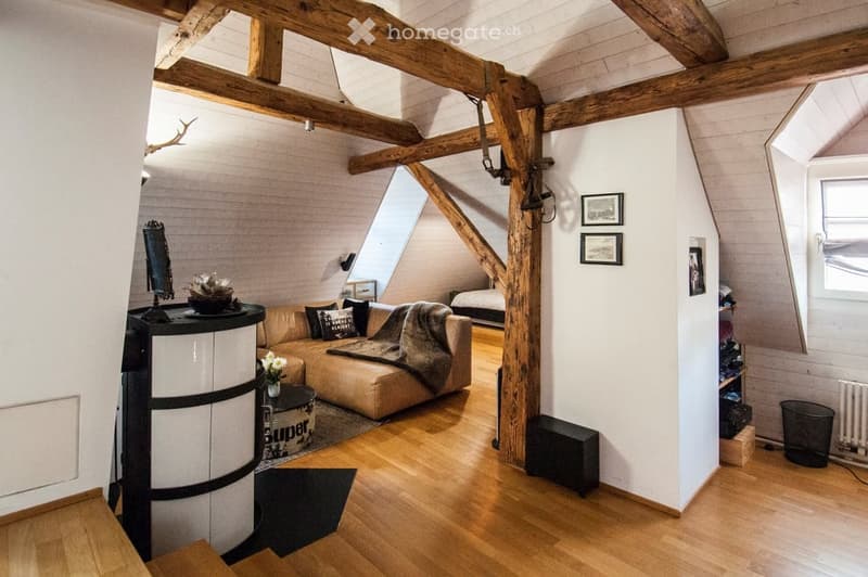 A cozy attic living room with exposed wooden beams, a leather sofa, a coffee table, a mirror, picture frames, and a small bed in the background.