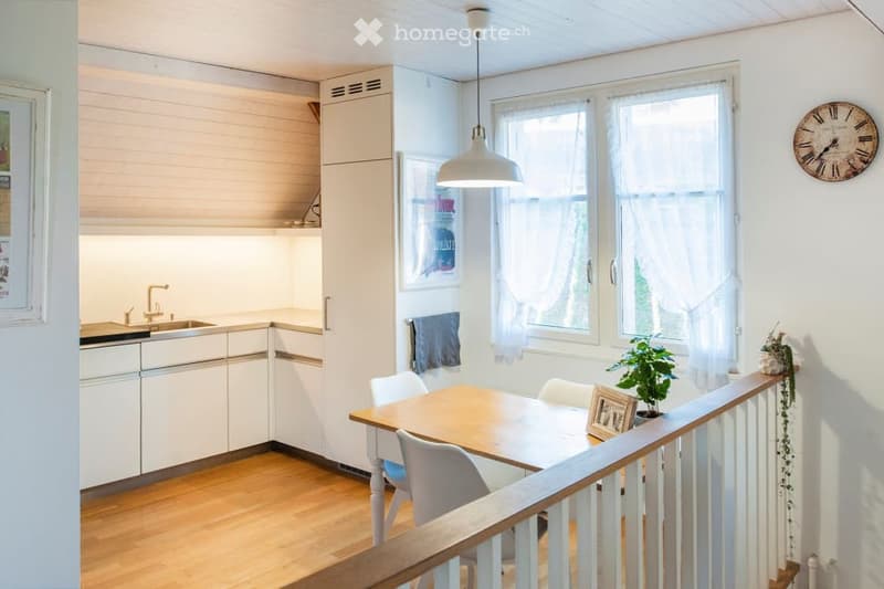 Bright, modern kitchen and dining area with white cabinets, a wooden dining table, white chairs, a pendant light, and a clock on the wall. The space features large windows with white curtains and a wooden floor.