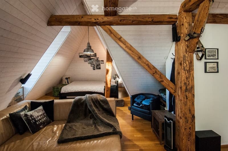 Loft-style living space with exposed wooden beams, a sofa, armchair, bed, small table, bookshelf, and picture frames on the wall.