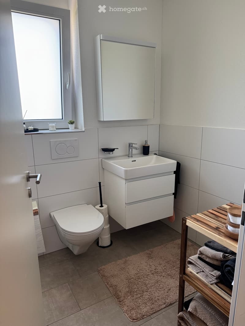 Modern bathroom with a toilet, sink, mirror, and a wooden shelf holding towels. The room features a beige tiled floor and walls, a window with a small plant on the sill, and a towel rack with a folded towel.