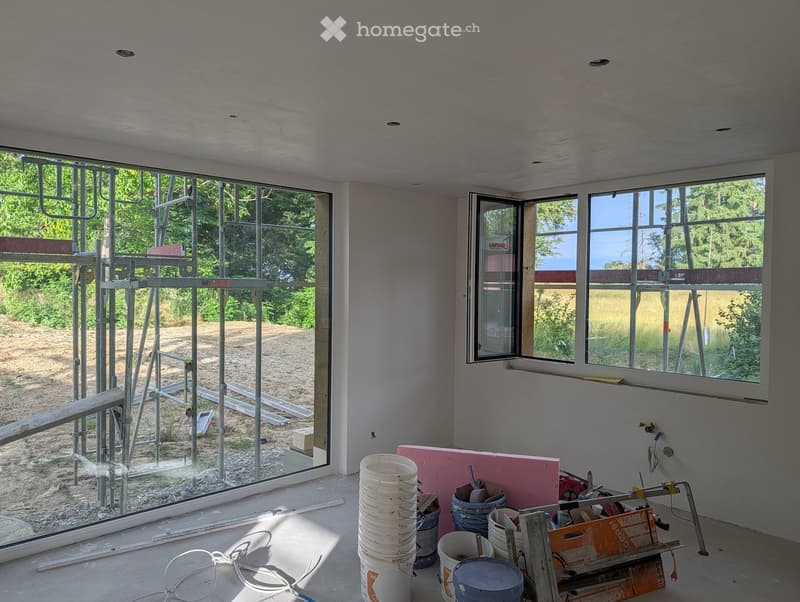 Empty room with unfinished walls and floor, large windows, and construction materials scattered around.