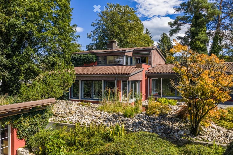 A modern, two-story house with red brick exterior and large windows, surrounded by lush greenery and a landscaped garden with rocks and shrubs.