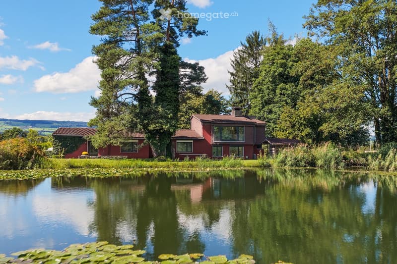 A red brick house with a tiled roof, surrounded by lush greenery and a pond in the foreground.