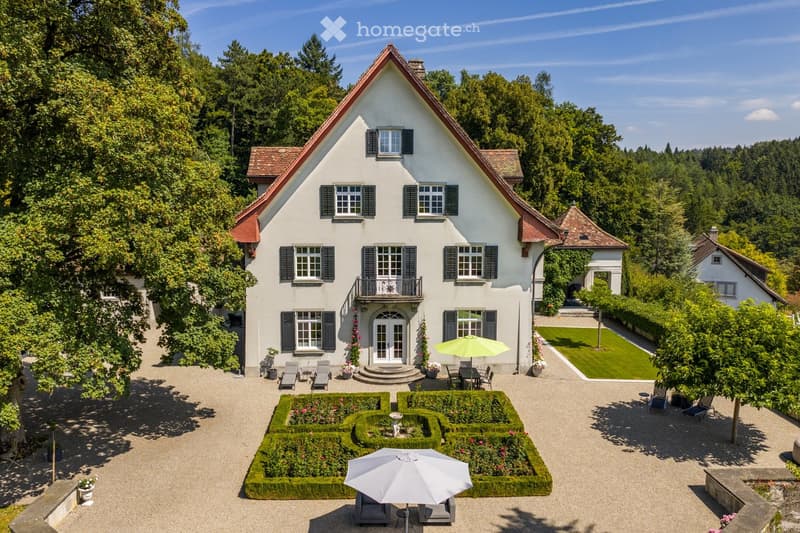 A large, two-story white house with a red-tiled roof and black shutters, surrounded by a well-maintained garden and outdoor seating areas.