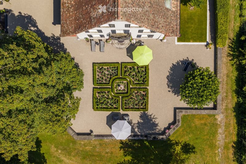 Aerial view of a well-maintained garden with a central circular pattern, surrounded by neatly trimmed hedges and flower beds. The area includes outdoor seating with umbrellas, a small pond, and a white building in the background.