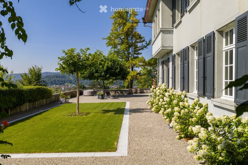 Well-maintained garden with a neatly trimmed lawn, gravel pathway, and surrounding trees and shrubs. The house features classic architecture with shutters and a view of the landscape.