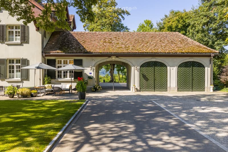 A two-story house with a stone facade, tiled roof, and large green garage doors. The front yard features a well-maintained lawn, outdoor seating area with umbrellas, and potted plants.