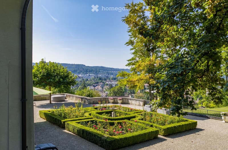 Well-maintained garden with neatly trimmed hedges, flower beds, and a scenic city view in the background.