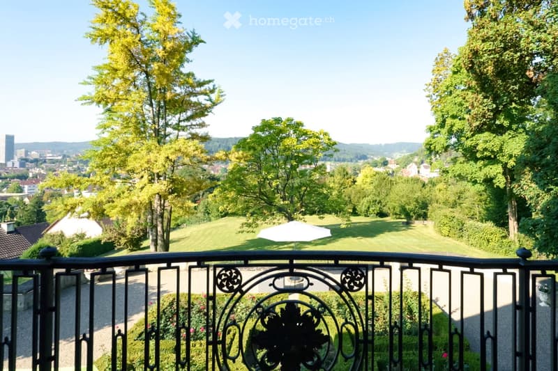 Balcony with ornate metal railing overlooking a landscaped garden and cityscape, featuring trees, a lawn, and distant buildings.