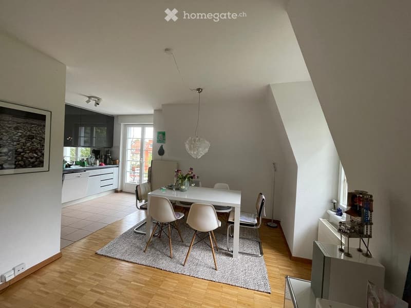 Bright, open-plan living and dining area with modern furniture, including a white dining table with chairs, a kitchen area with cabinets and appliances, and decorative elements like a framed picture and a vase with flowers.