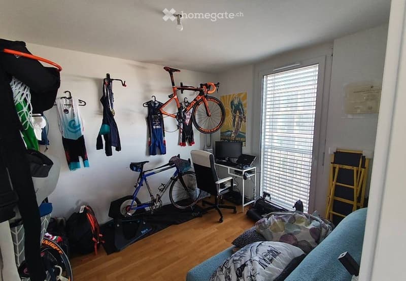 A furnished room with a desk, chair, laptop, bicycles, and various cycling gear. The room features wooden flooring, a window with blinds, and a bed with a blue blanket.