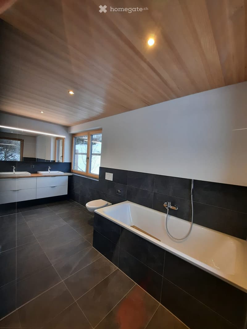 Modern bathroom with dark tiled walls and floor, white bathtub, toilet, double sink, and large mirror. Features wooden slatted ceiling and large window.