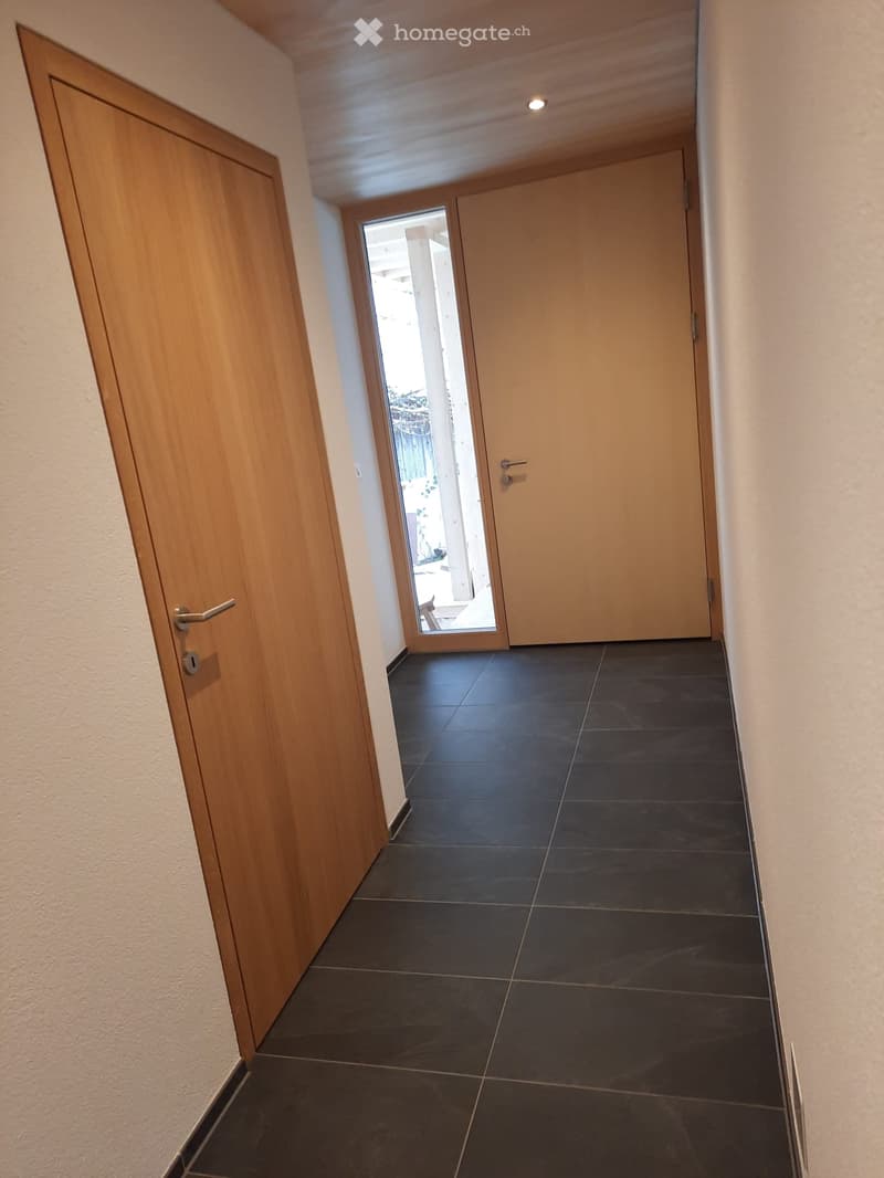 A narrow hallway with dark tiled flooring, light-colored walls, and wooden doors on both sides. A sliding glass door at the end provides a view to an outdoor area.
