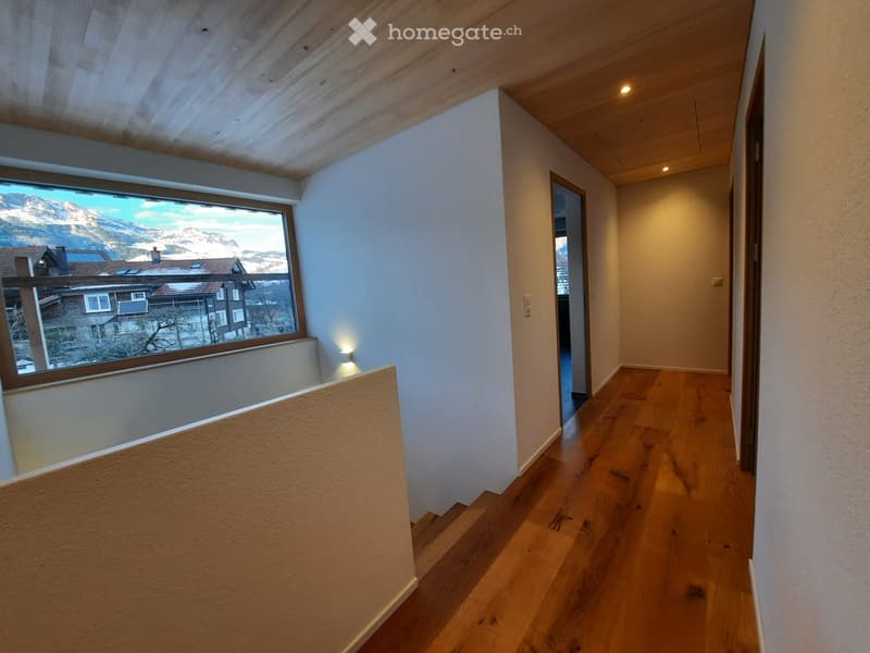 A narrow hallway with wooden flooring, white walls, and recessed lighting. A large window on the left offers a scenic mountain view.