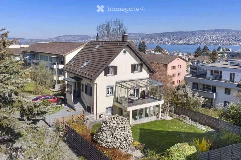 A two-story house with a modern design, featuring a large glass balcony, surrounded by greenery and overlooking a scenic lake and distant mountains.