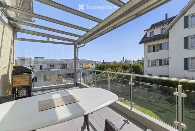 A modern glass-enclosed balcony with a white table and a view of a suburban neighborhood.