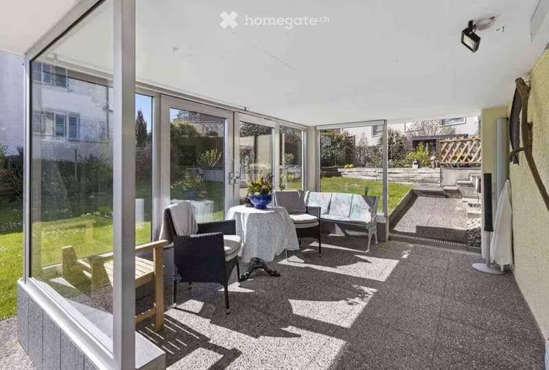 Bright, enclosed patio with large glass walls, carpeted floor, sofa, table, chairs, and a flower pot.