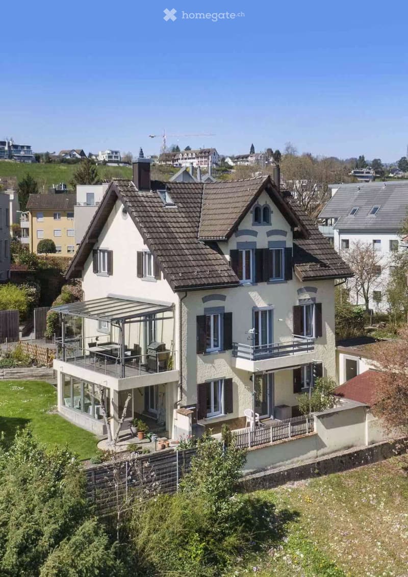 Two-story house with a tiled roof, multiple windows, and a balcony. The house features a covered patio area with outdoor furniture and a small garden with potted plants.