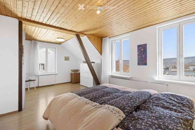 Bright bedroom with wooden floors, slanted wooden ceiling, large windows, bed, small table, chair, and artwork on the walls.