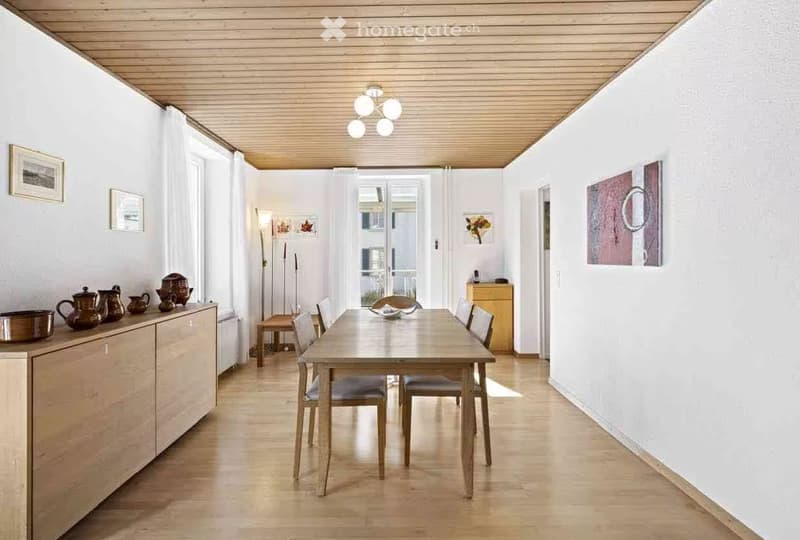 Bright dining area with wooden table and chairs, cabinet with teapots, and artwork on walls.