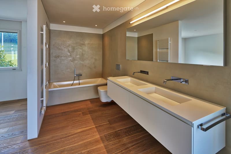 Modern bathroom with a large bathtub, double sink vanity, sleek faucets, and a large mirror above the sink. The room features wooden flooring and neutral-colored walls.