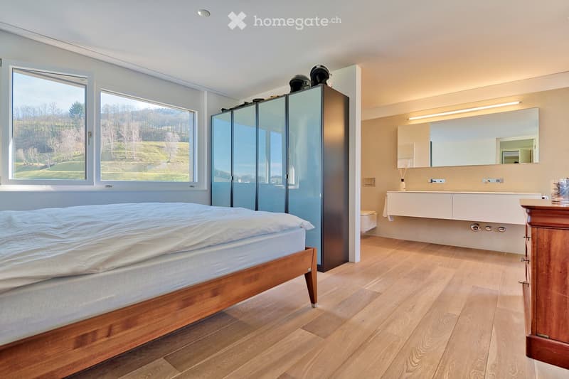 Spacious bedroom with large windows offering a scenic view, featuring a wooden bed, modern wardrobe, and an en-suite bathroom with a sink and mirror.