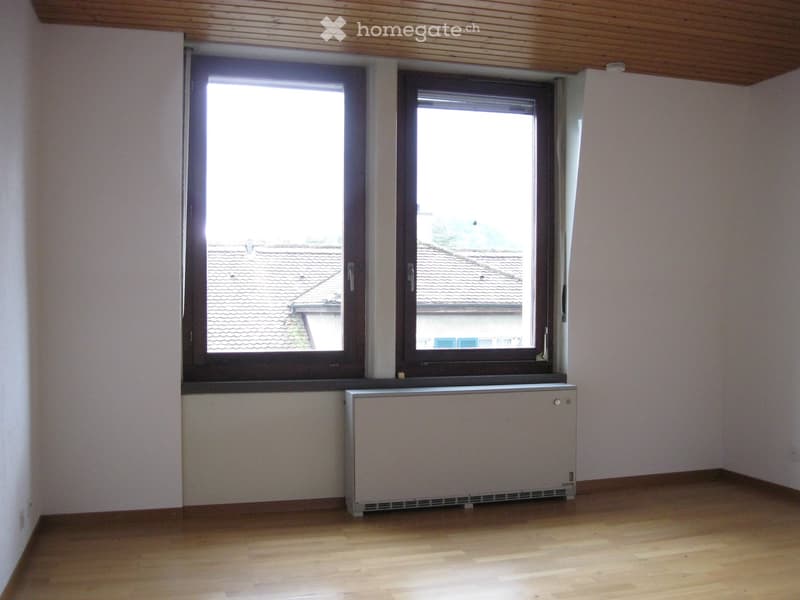 Empty room with wooden flooring, white walls, and two windows overlooking a tiled roof. A radiator is placed beneath the windows.