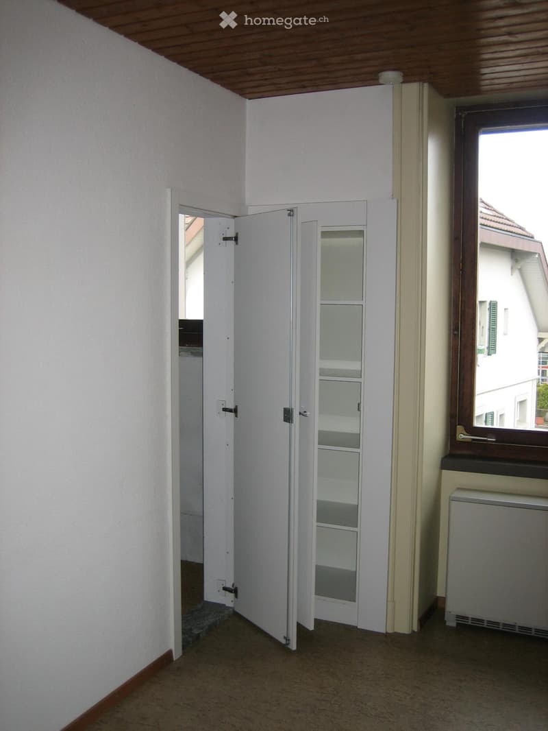 Empty room with white walls, wooden ceiling, tiled floor, and a window with wooden frame. A door leads to another room.