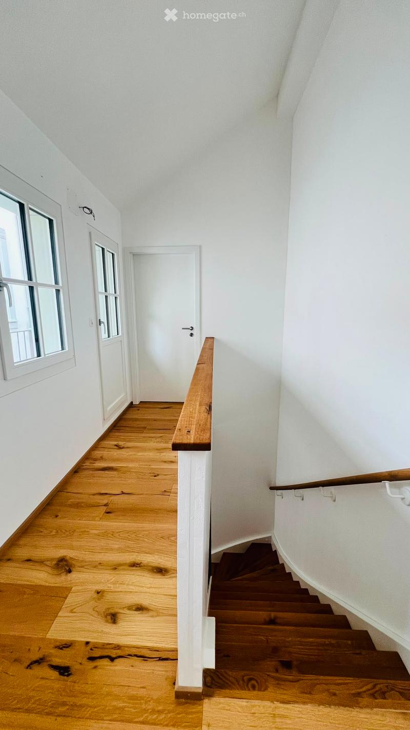 A narrow hallway with wooden flooring, white walls, and a staircase leading upwards. Features include a wooden handrail, a white door, and a window with multiple panes on the left side.