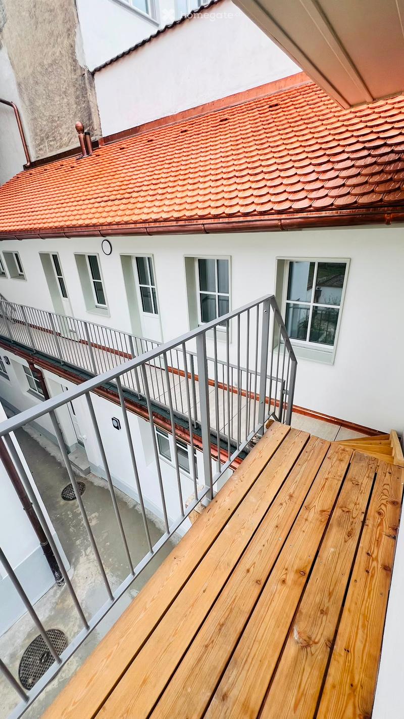 Balcony with wooden decking, metal railing, overlooking a building with white walls, multiple windows, and a red-tiled roof.
