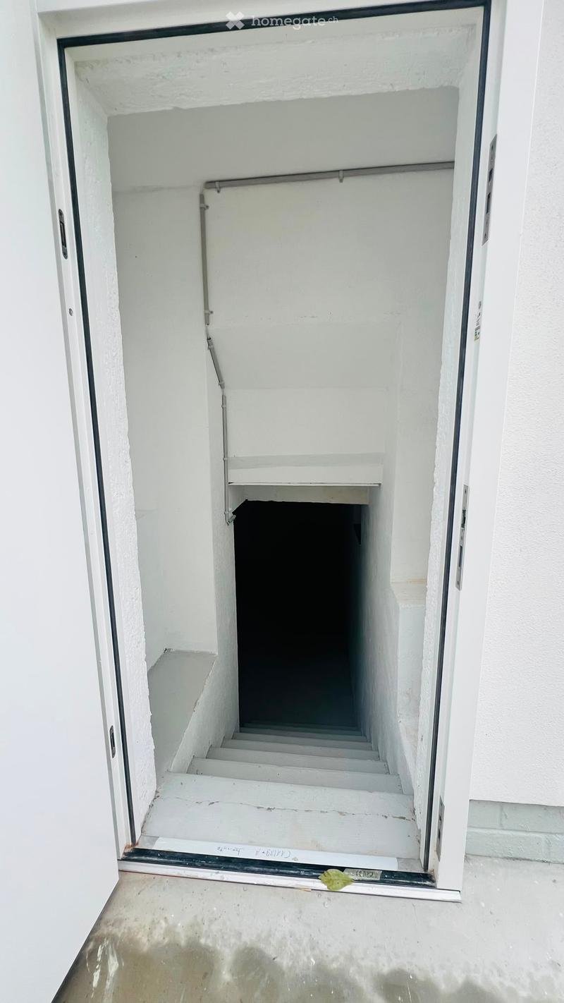 A narrow, white-walled stairwell leading downwards with a metal railing and a small shelf at the top.