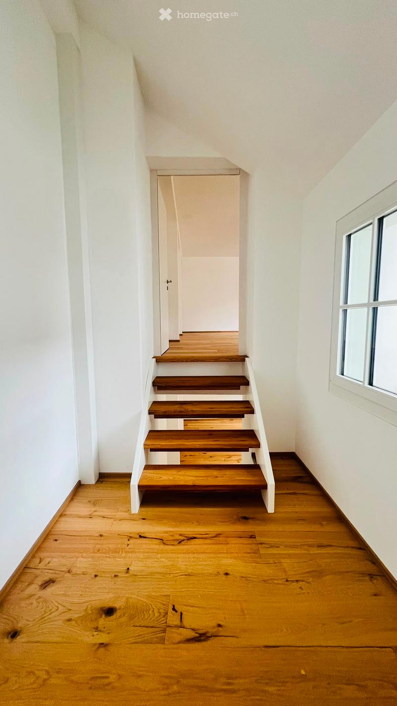 A narrow hallway with wooden flooring and white walls, featuring a staircase with wooden steps and white railings leading upwards.