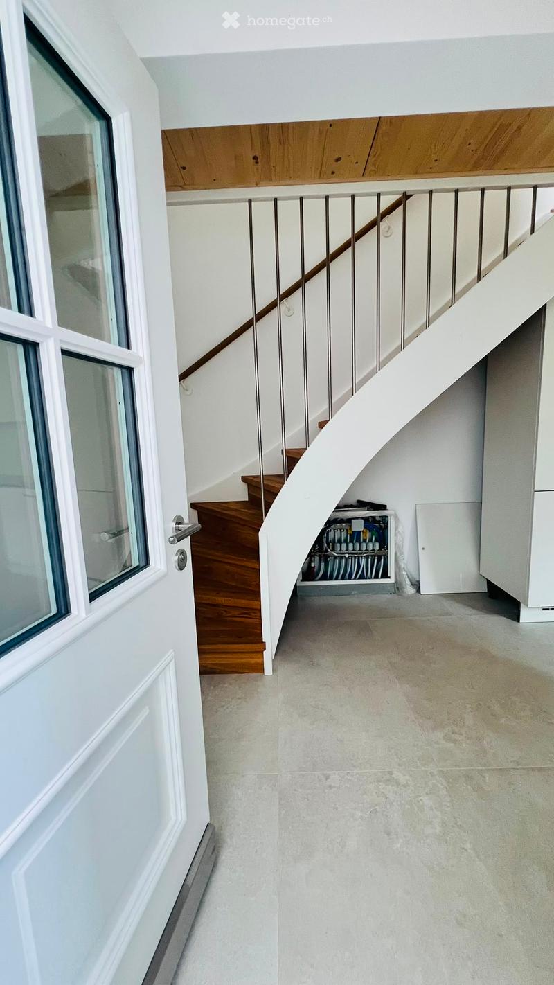 Modern hallway with a staircase featuring wooden steps and a metal railing, white walls, and a grey tiled floor. A window is visible on the left side, and a small utility area is visible under the stairs.