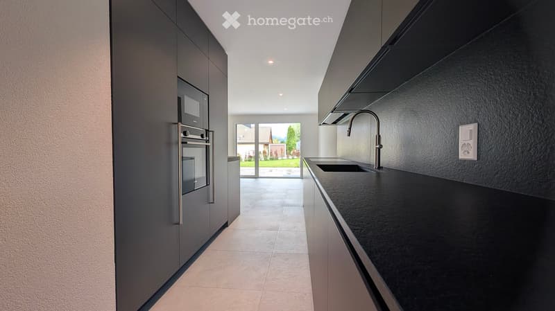 Modern kitchen with dark grey cabinets, black countertop, stainless steel sink and faucet, and built-in oven. Large window offers a view of the garden.