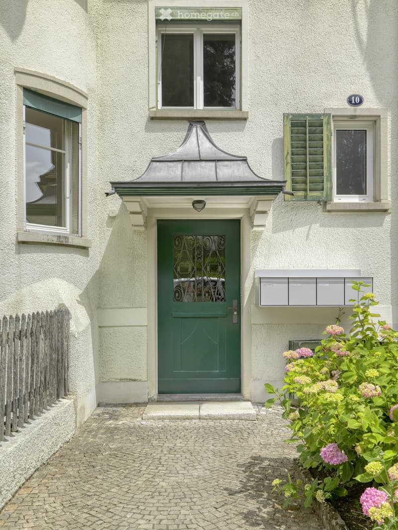 A two-story building with a green front door, decorative metalwork, and a small garden with pink flowers. The entrance features a stone step and a cobblestone pathway.
