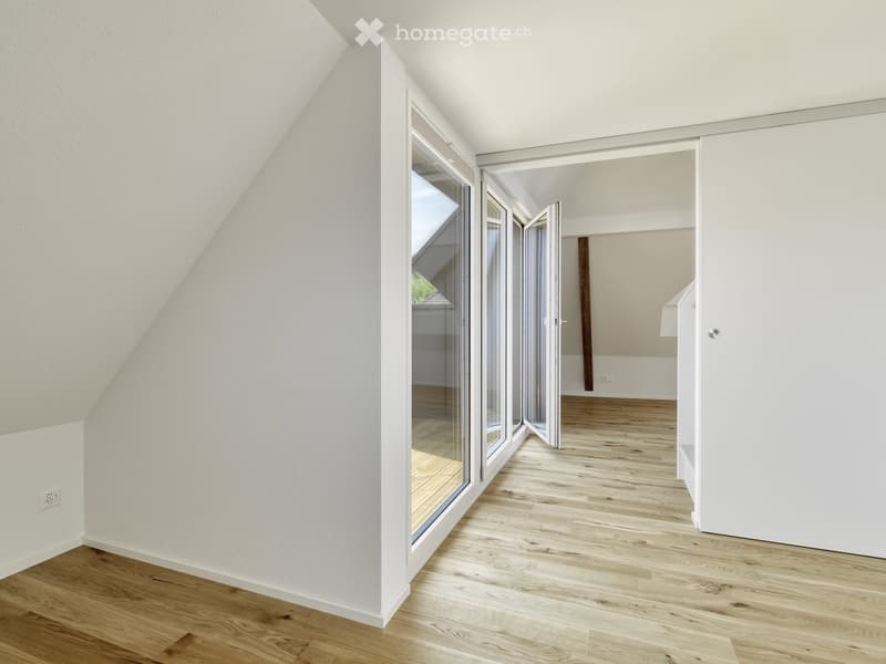 Empty attic space with wooden flooring, white walls, large windows, and open sliding doors leading to another room.