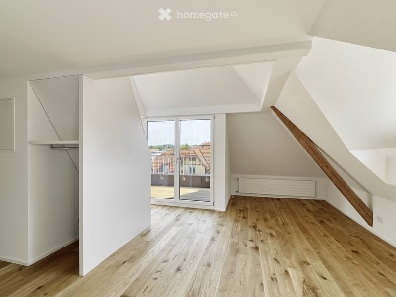 Bright, empty attic room with wooden flooring, white walls, and a large skylight window. Features a built-in closet and a staircase leading upwards.