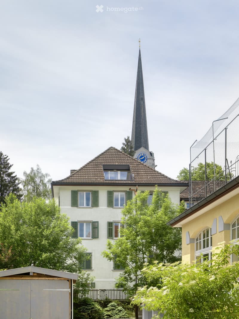 A two-story residential building with a steeply pitched roof, green shutters, and a prominent clock tower with a tall spire. The building is surrounded by lush greenery and trees.