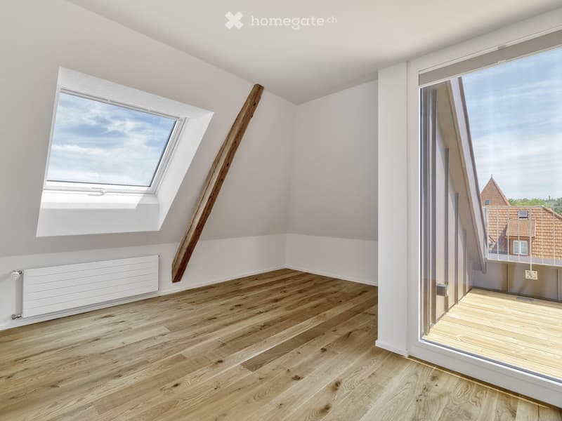 Bright, empty attic room with wooden flooring, white walls, large skylight window, and a sliding glass door leading to a balcony.